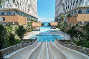 Piscina de resort com escorregador aquático, palmeiras e vista para o mar.
