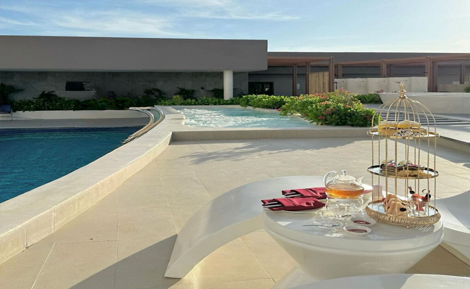 Un área de comedor junto a la piscina, con un soporte de tres niveles para postres, una mesa con tetera y tazas, y vistas a la piscina y al edificio al fondo.