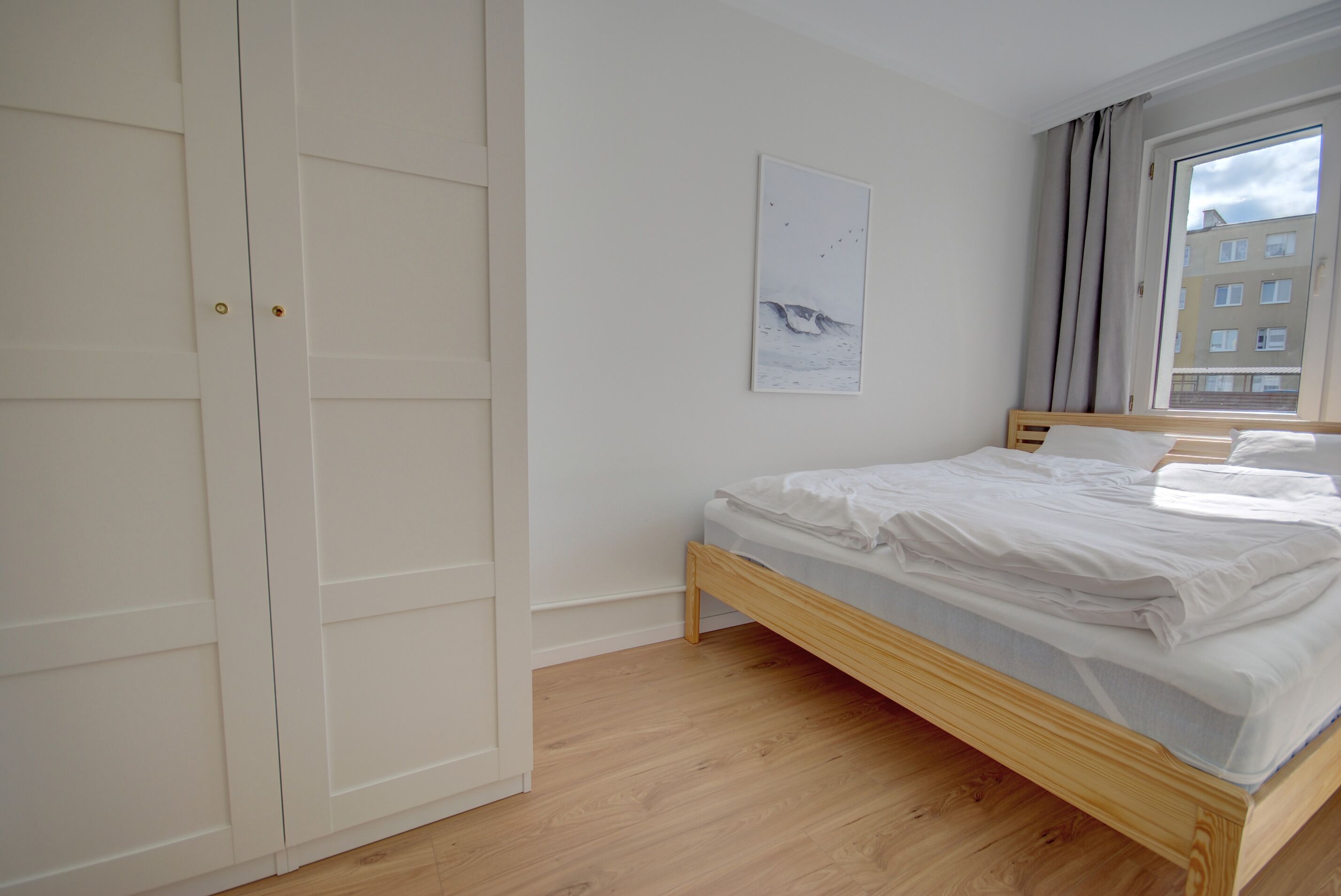 Un dormitorio con una cama de madera, ropa de cama blanca, un armario blanco y una ventana con cortinas.