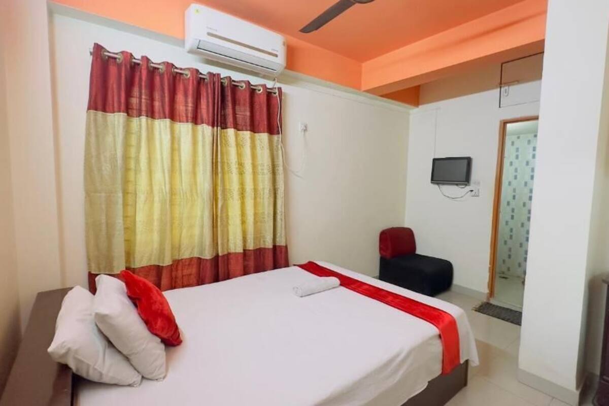 A hotel room with a bed, a red and yellow curtain, an air conditioning unit, a television, and a small desk.