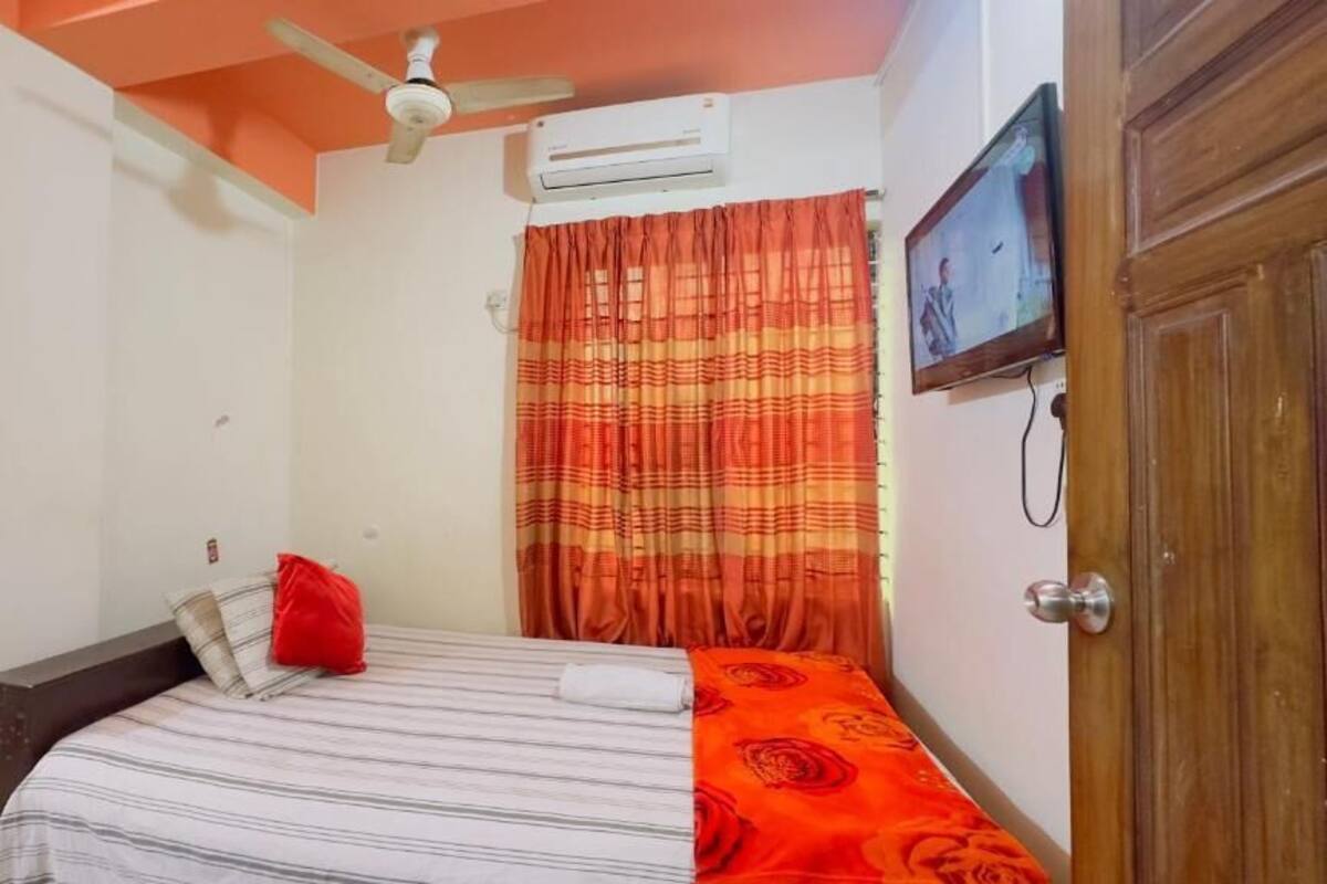 A bedroom with a bed, a ceiling fan, an air conditioner, a television, and a door.