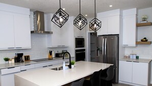 A modern kitchen with white cabinetry, a central island with bar stools, stainless steel appliances, and geometric pendant lighting.