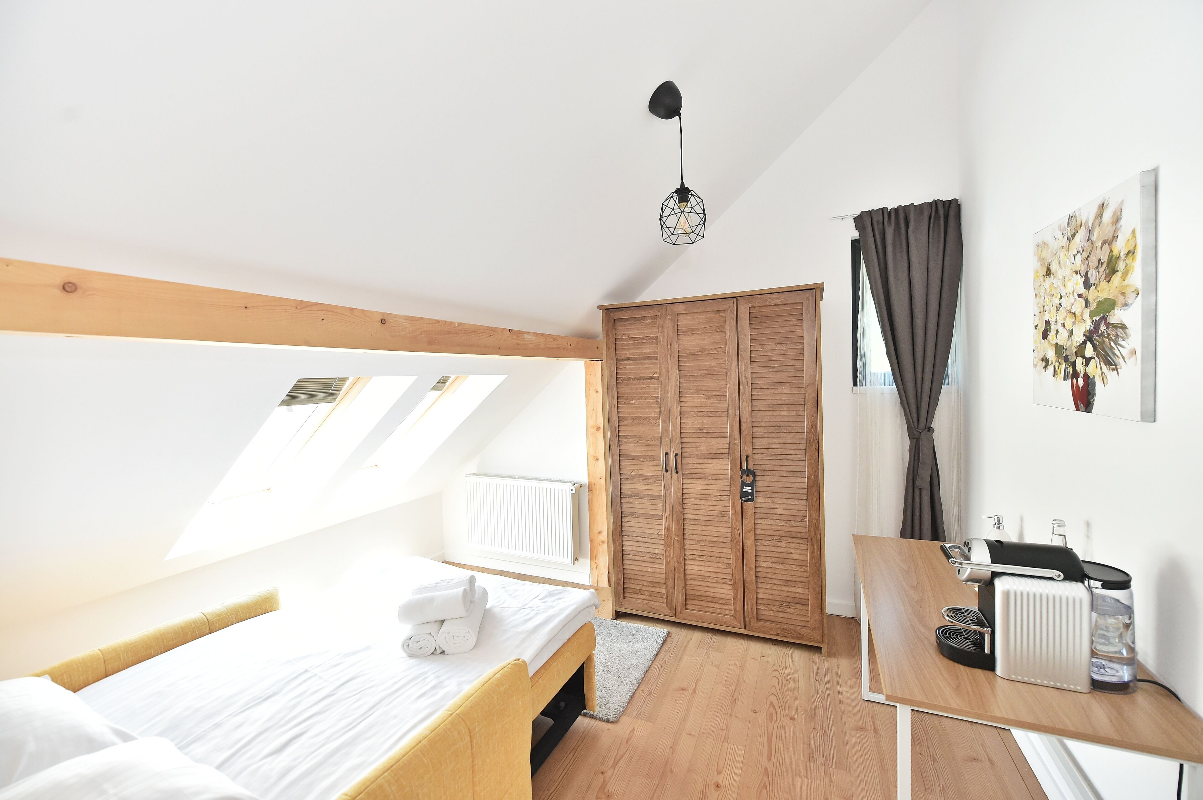A bedroom with a bed, a wooden wardrobe, a desk with a radiator, and a ceiling light.