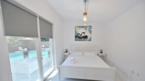 A bedroom with a large bed, a nightstand, a painting of swans, and a view of an outdoor swimming pool.
