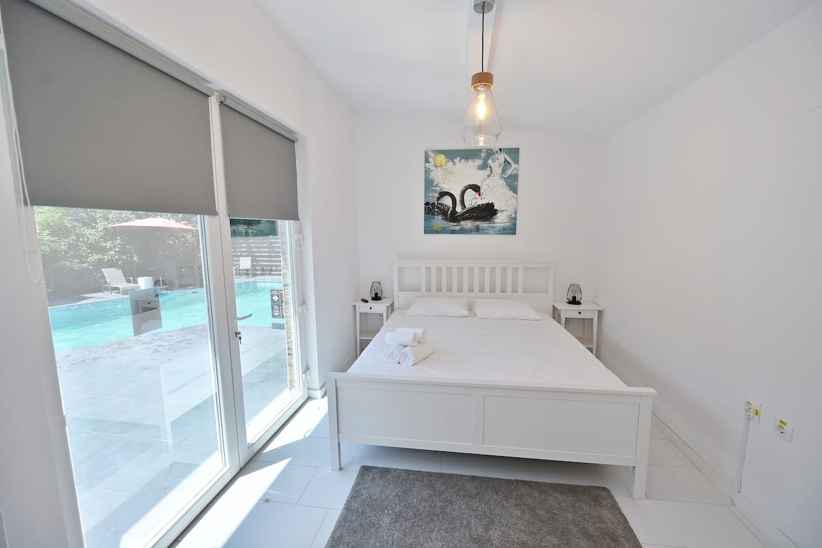A bedroom with a large bed, a nightstand, a painting of swans, and a view of an outdoor swimming pool.