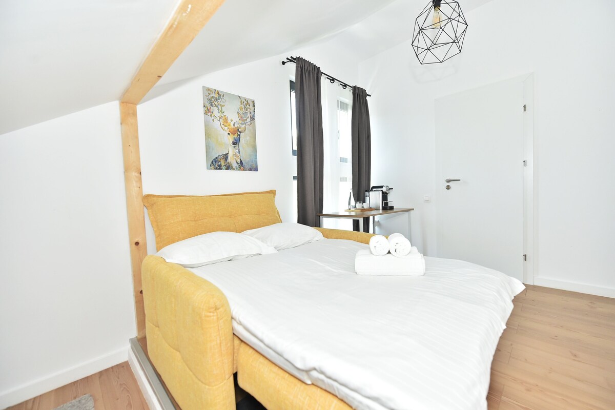 A bedroom with a bed, a painting, a window with curtains, a bedside table, and a door.