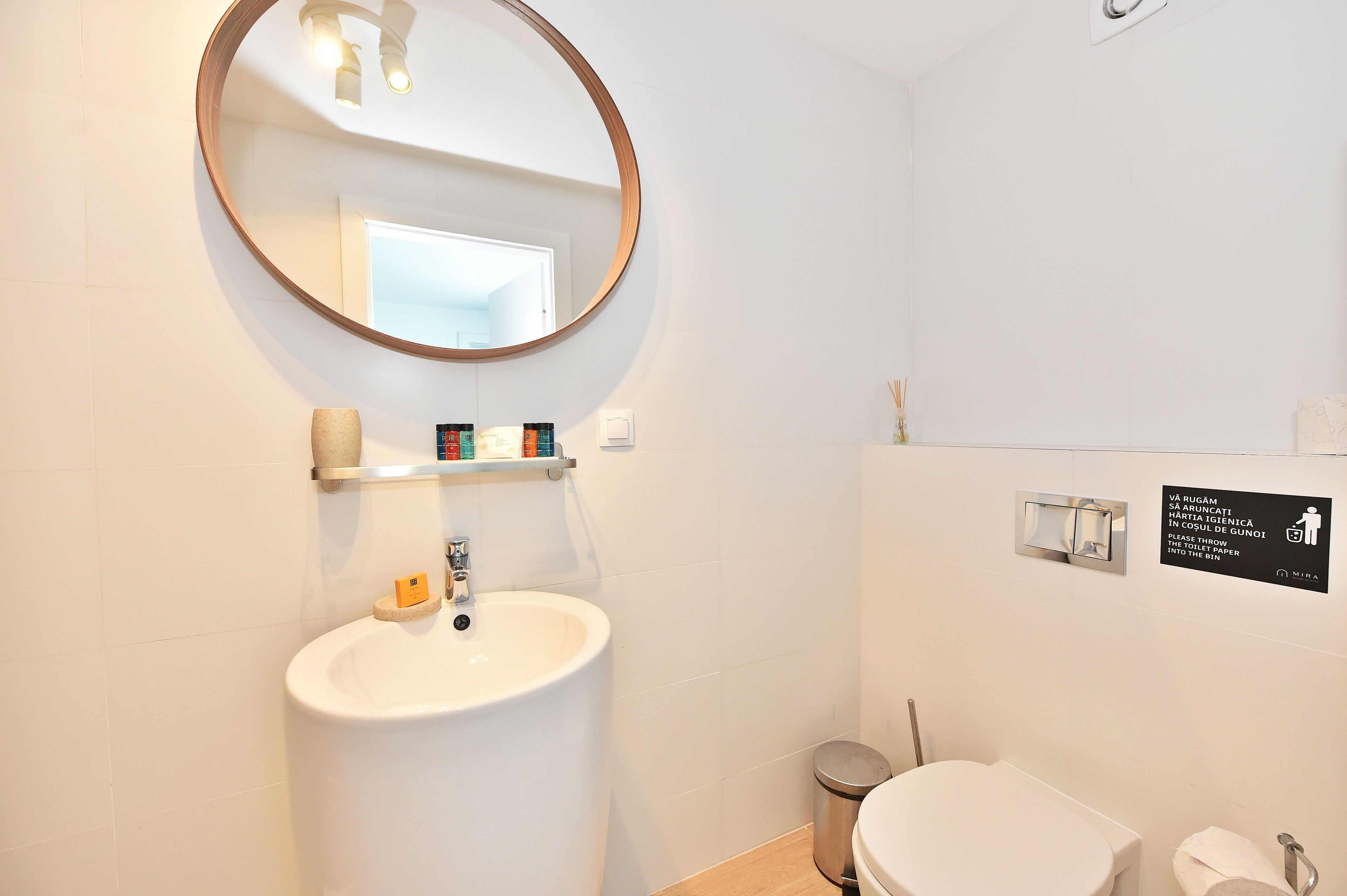 Ein modernes Badezimmer mit einem runden Spiegel, einem Waschbecken, einer Toilette und einem Mülleimer. An der Wand befindet sich ein Schild mit Anweisungen.