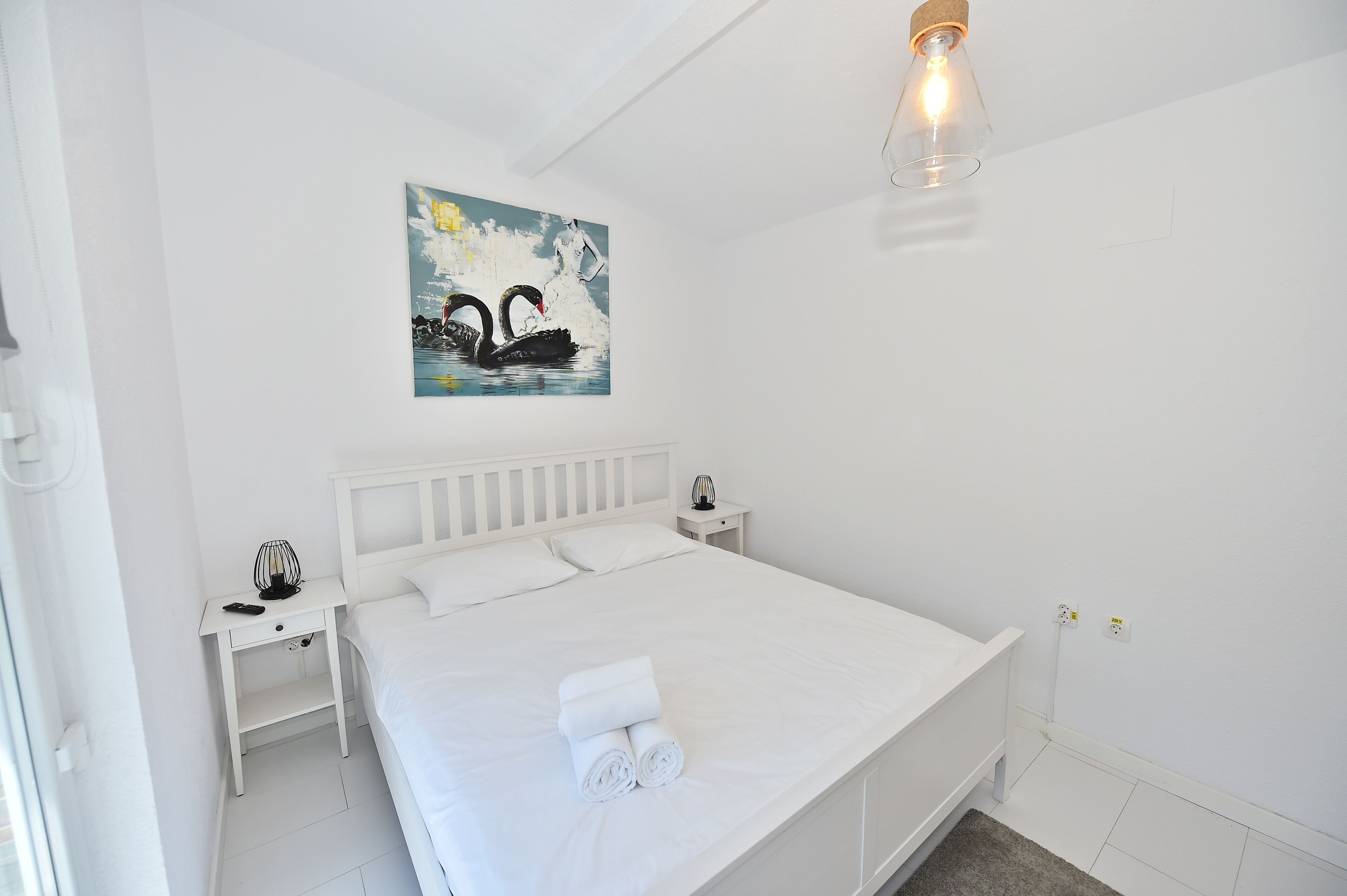 Une chambre à coucher blanche, avec un lit, une table de chevet, une lampe fixée au mur et un tableau représentant des cygnes.