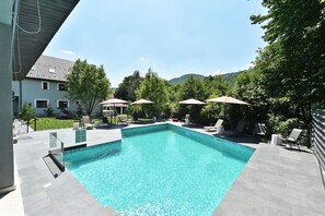 A rectangular swimming pool with clear blue water, surrounded by a tiled deck. There are lounge chairs and umbrellas, and a building with a tiled roof in the background. - Mira by Atra (Valea Doftanei)