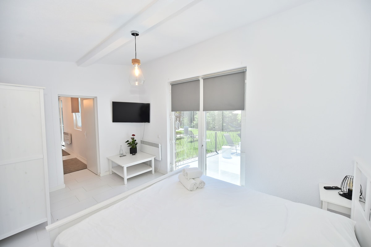 A modern bedroom with a large bed, a TV mounted on the wall, and a balcony with a view.