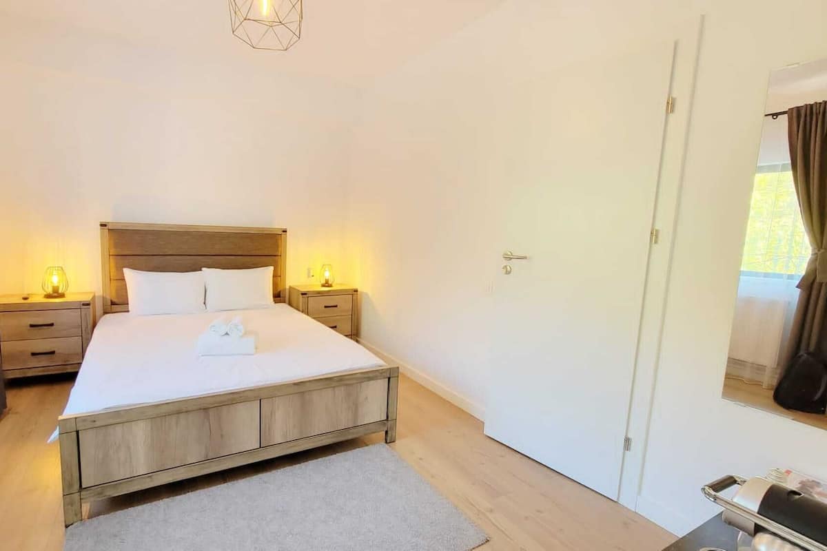 A bedroom with a wooden bed, white bedding, two bedside tables with lamps, a door, and a window with curtains.