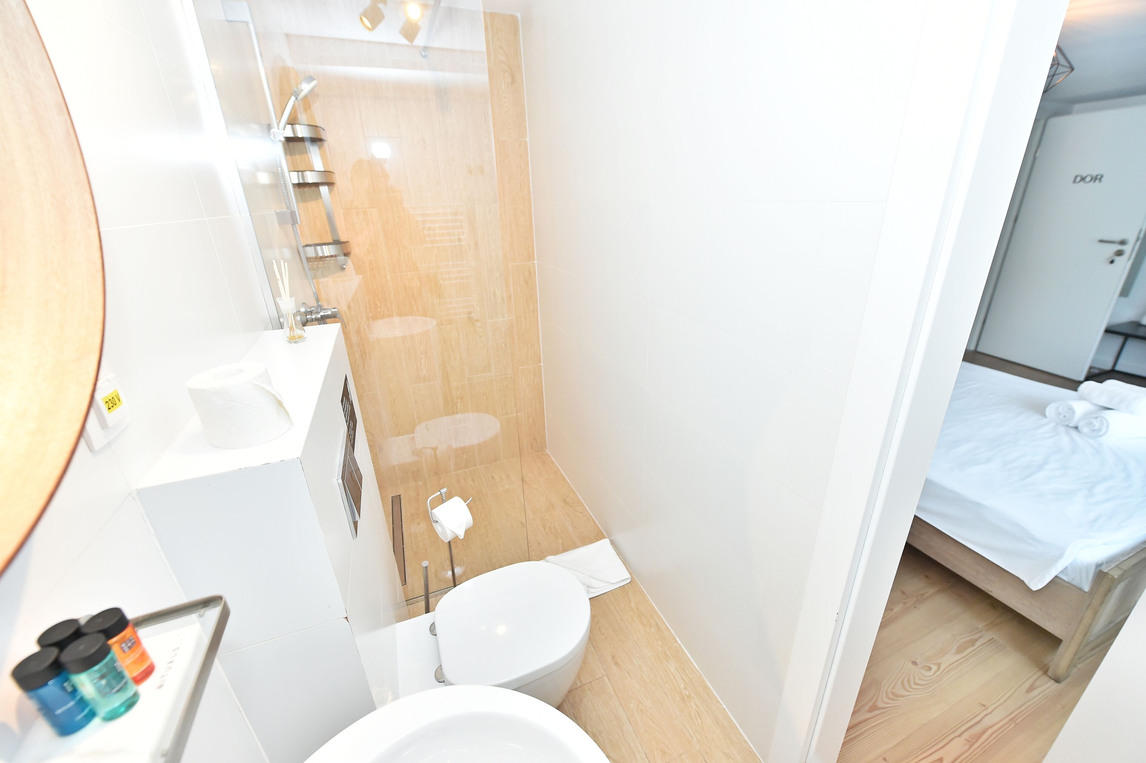 A compact bathroom with a toilet, sink, and shower. There is a small shelf with toiletries and a towel rack.
