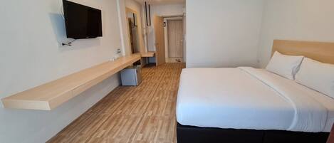 A hotel room with a bed, a TV mounted on the wall, a wooden floor, and a small desk with a chair.