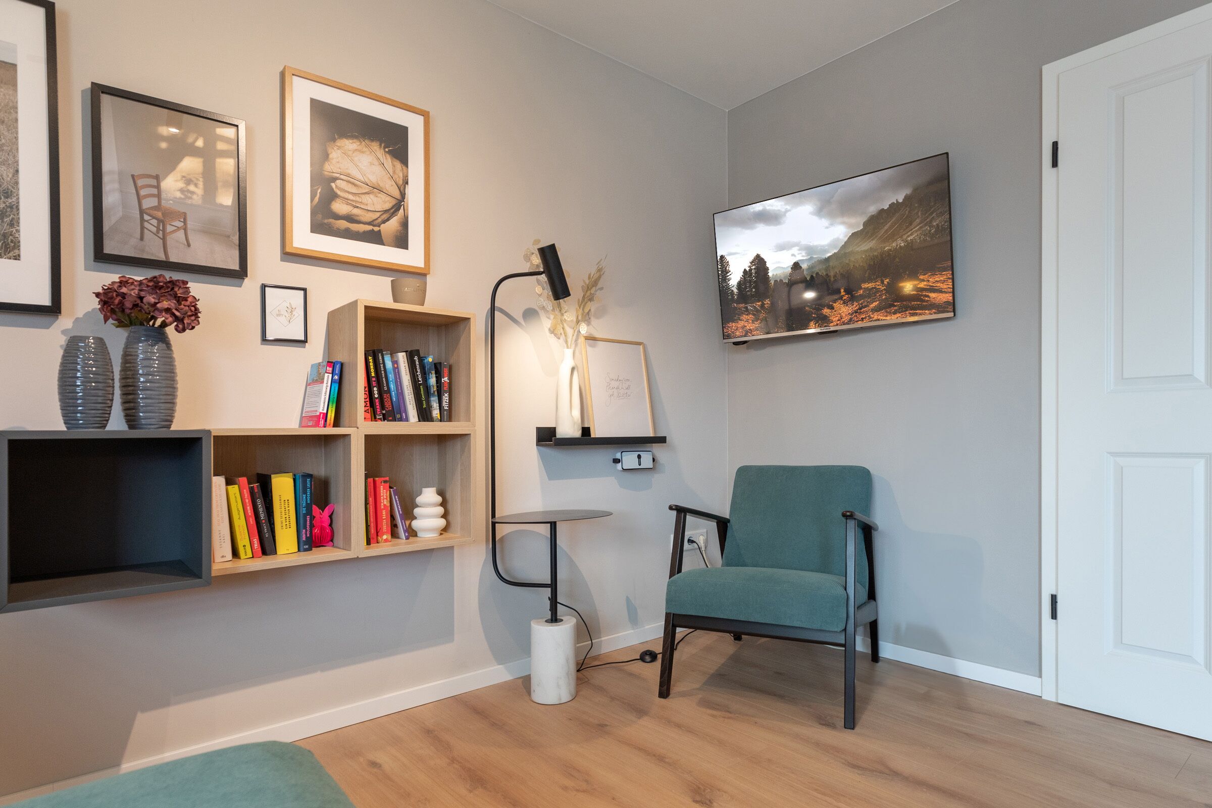 A modern living room with a teal armchair, wooden bookshelves, a small table, and framed artwork on the walls.
