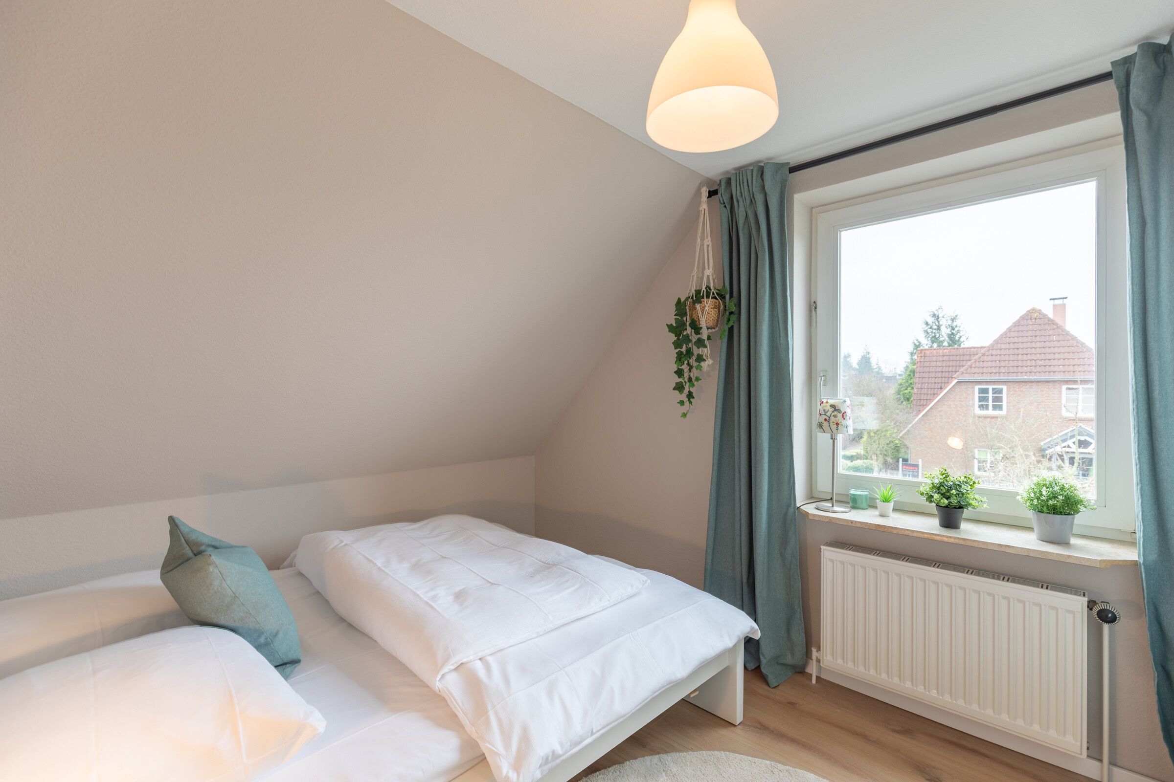 A bedroom with two beds, a window with curtains, a hanging plant, and a radiator.