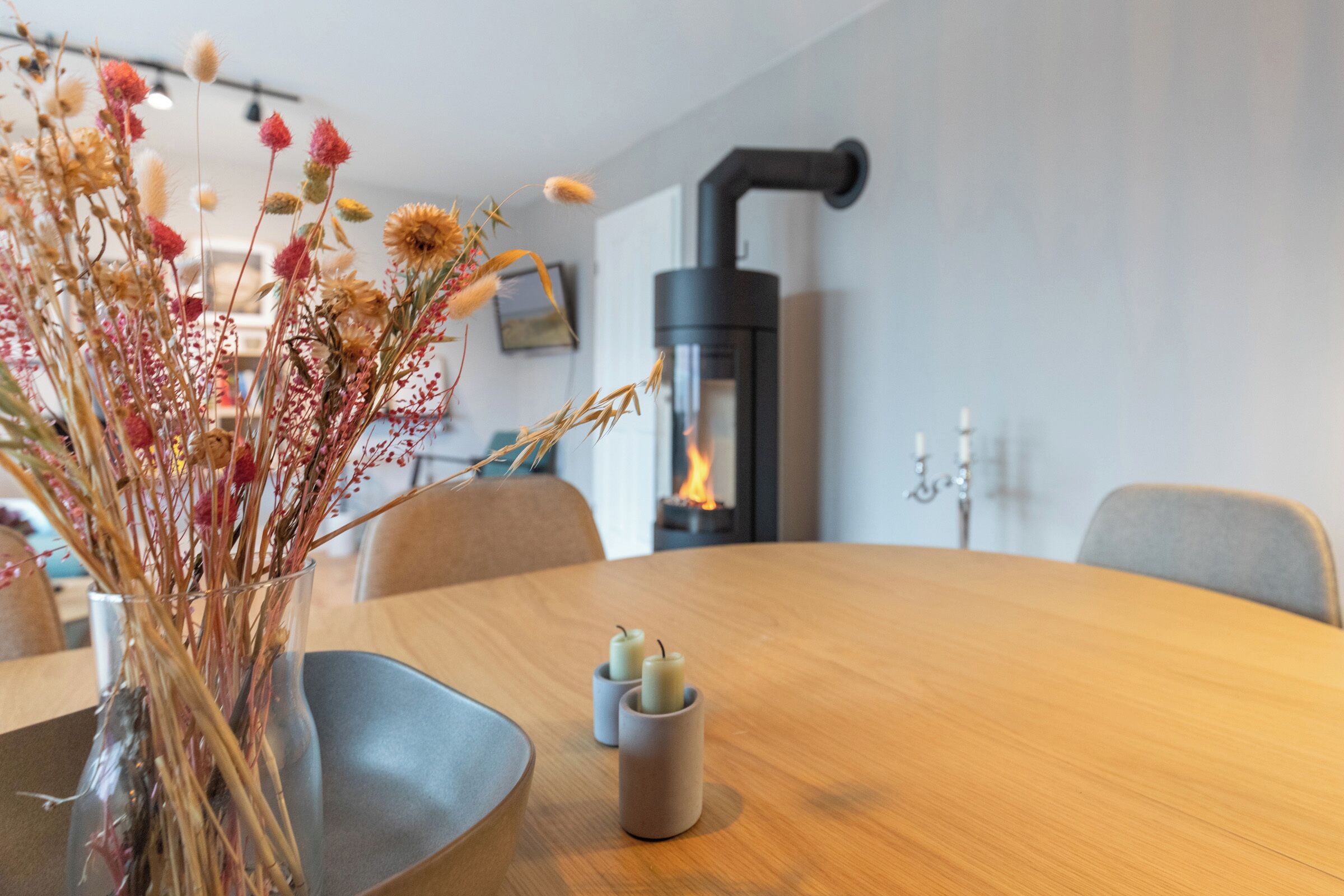 Et spisebord med stearinlys, en vase med visne blomster og en brændeovn i baggrunden.