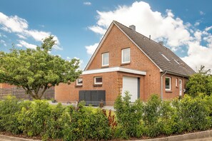 Une maison à deux étages, construite en briques, avec un toit en pente, une porte de garage et une cour clôturée agrémentée de divers arbustes.