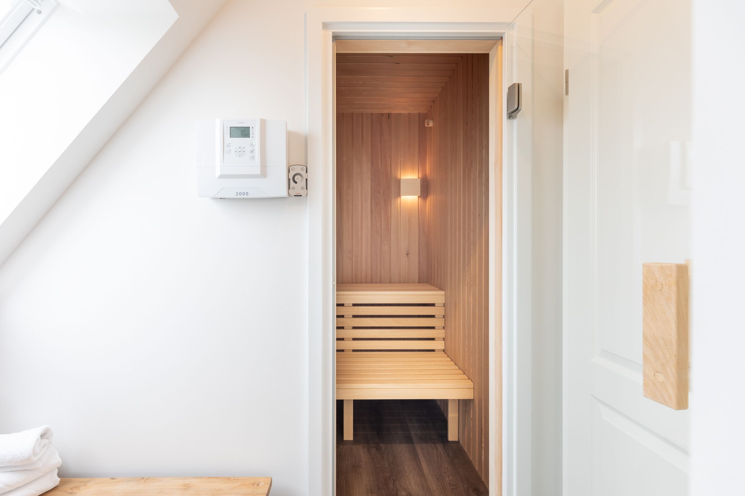 Sala de sauna com banco de madeira, painel de controle na parede e uma pequena pilha de toalhas.