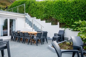 Une terrasse extérieure moderne, avec une table et des chaises en bois, un escalier menant à une porte vitrée et, en arrière-plan, une haie verdoyante et luxuriante.