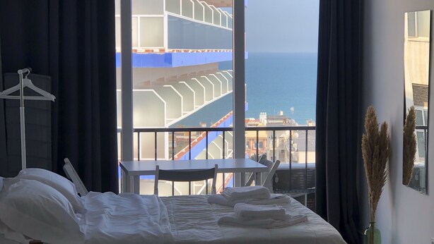 Habitación de hotel con una cama grande, una mesita y vistas al mar y un edificio con balcones.