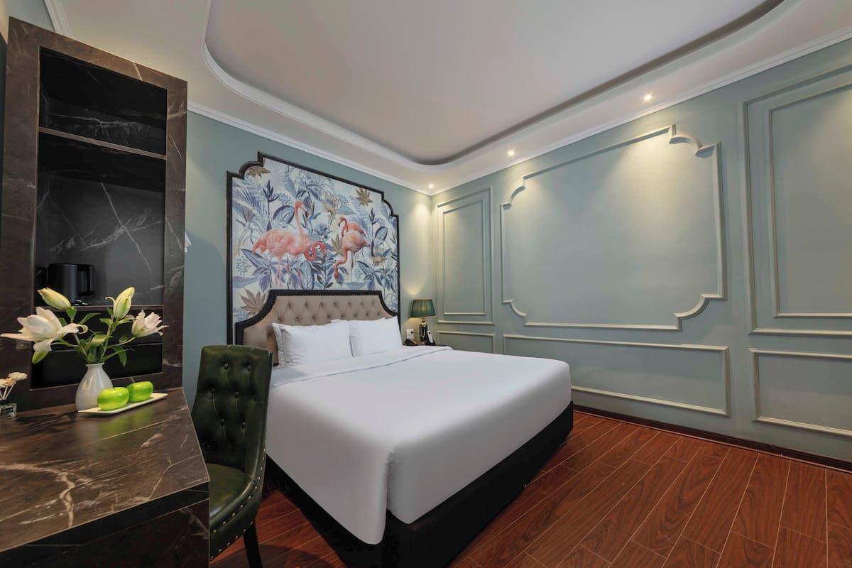 A hotel room with a large bed, a vanity with a mirror, a chair, a table with a vase of flowers, and a wall with a floral painting.