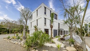 Un edificio moderno de dos pisos con fachada blanca, rodeado por un jardín ajardinado con diversas plantas y rocas.