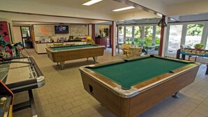 A pool area with two pool tables, a bar area with a television, and a view of the outdoors through large windows.
