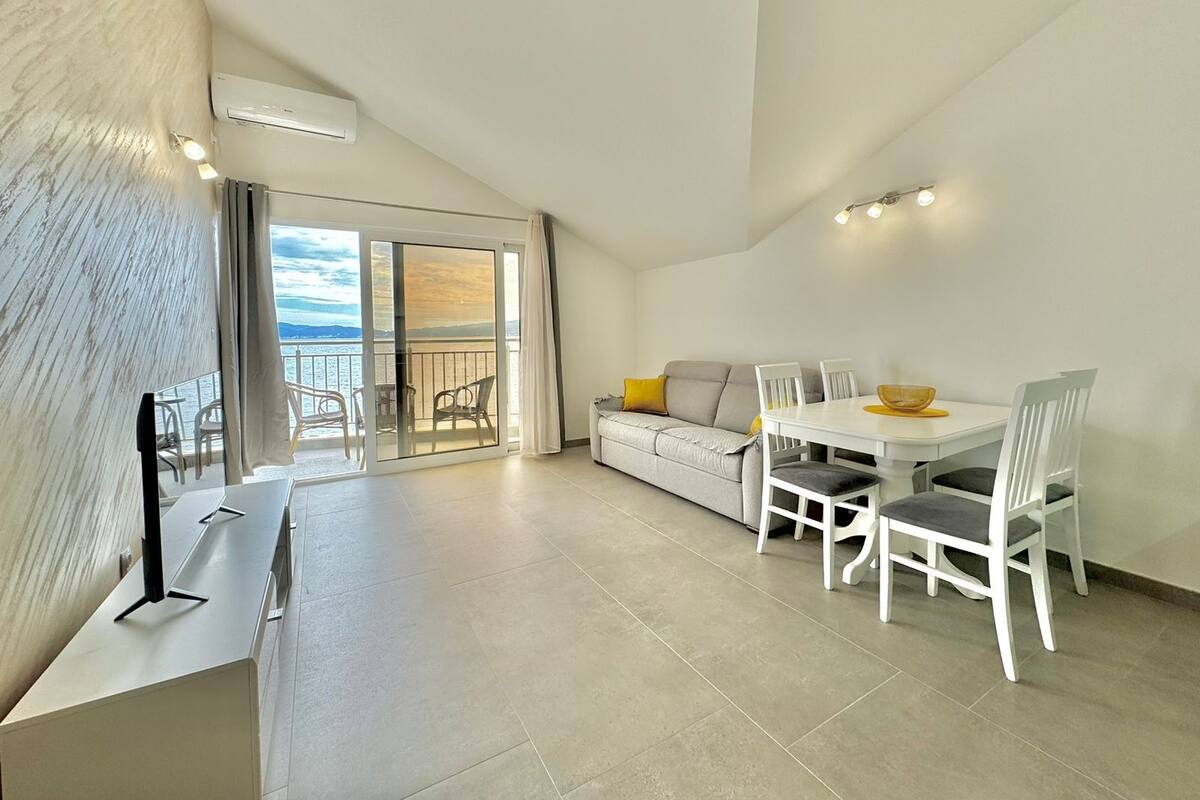 Ein modernes Wohnzimmer mit einer Couch, Esstisch und Stühlen. Eine Glastür führt auf einen Balkon mit Meerblick.
