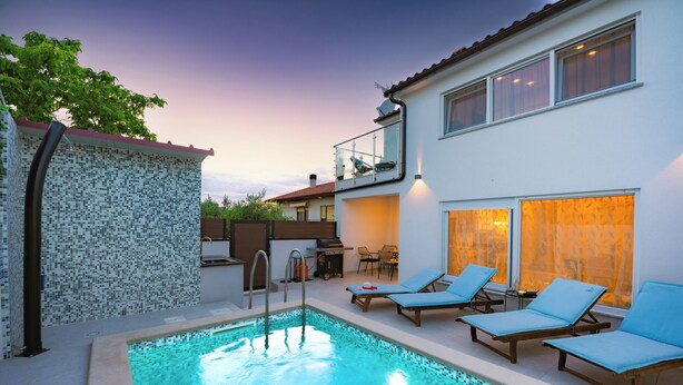 Une piscine avec des chaises longues, un espace barbecue et un bâtiment à deux étages doté d’un balcon.