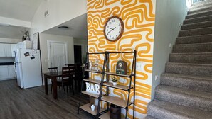 Une cuisine équipée d’un réfrigérateur, une table à manger avec des chaises, une horloge au mur et un escalier menant à l’étage.