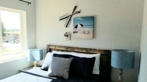 Een slaapkamer met een bed, een nachtkastje, een lamp, een raam en een aan de muur hangend schilderij.