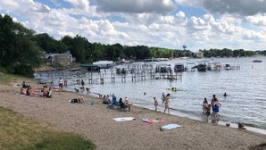 Een strand met mensen die zonnebaden en zwemmen, een steiger met boten, en een watertoren op de achtergrond.