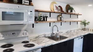 A modern kitchen with a white microwave, stove, and sink, wooden shelves with decorative items, and a marble countertop.