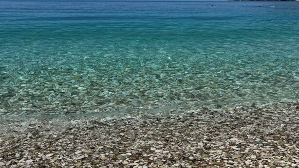 Visão clara de uma praia de seixos com águas calmas do mar e uma massa terrestre distante.