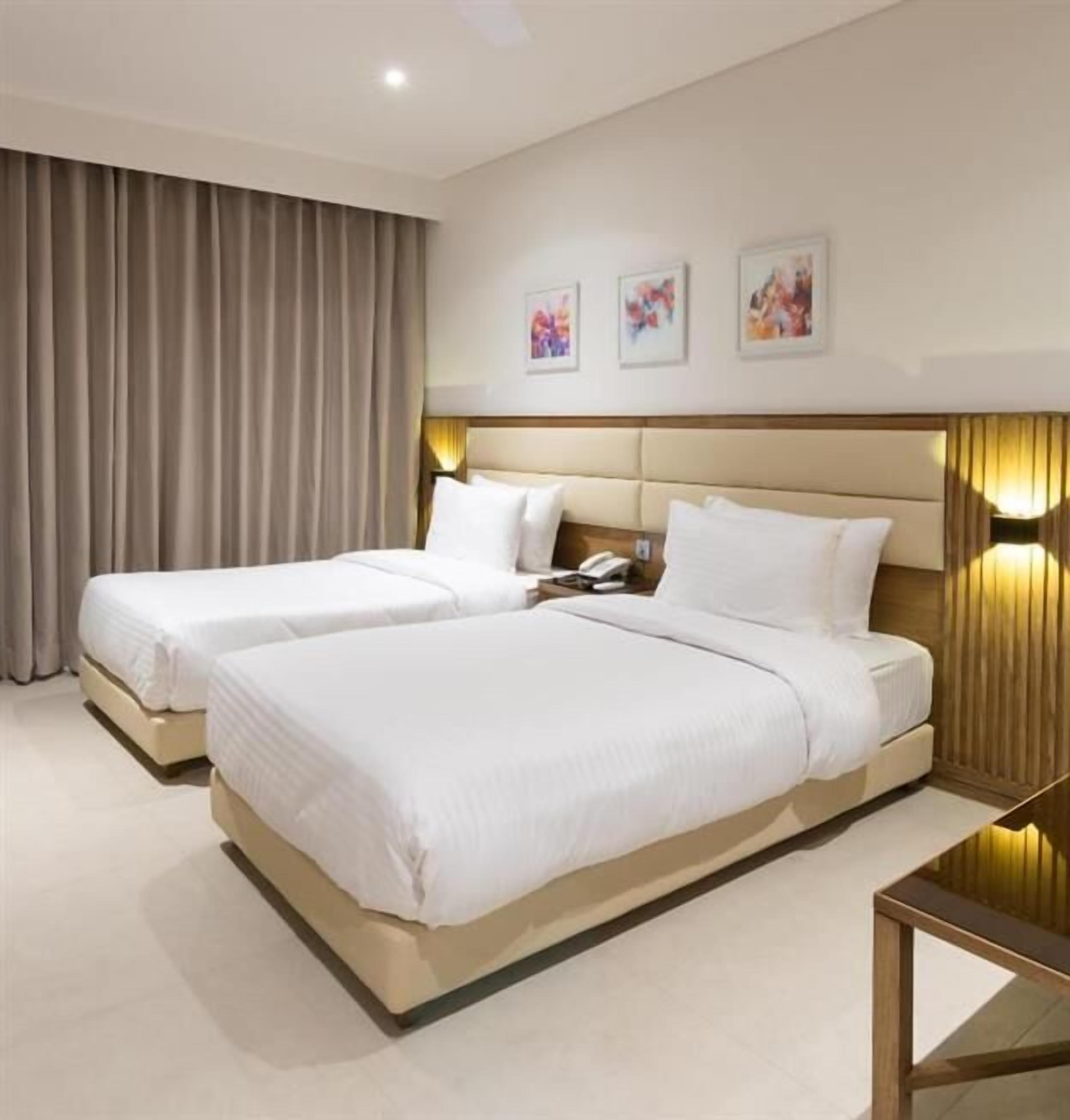 Habitación de hotel con dos camas, cabecero de madera, mesita de noche con lámpara y tres cuadros enmarcados en la pared.