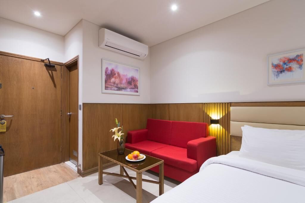 Habitación de hotel con puerta de madera, sofá rojo, una mesita con un florero y fruta, una cama y dos aires acondicionados instalados en la pared.