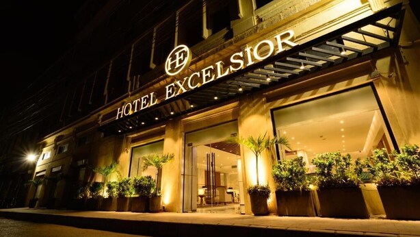 L’entrée d’un hôtel, avec une enseigne indiquant « HÔTEL EXCELSIOR » et une façade moderne agrémentée de plantes en pot.