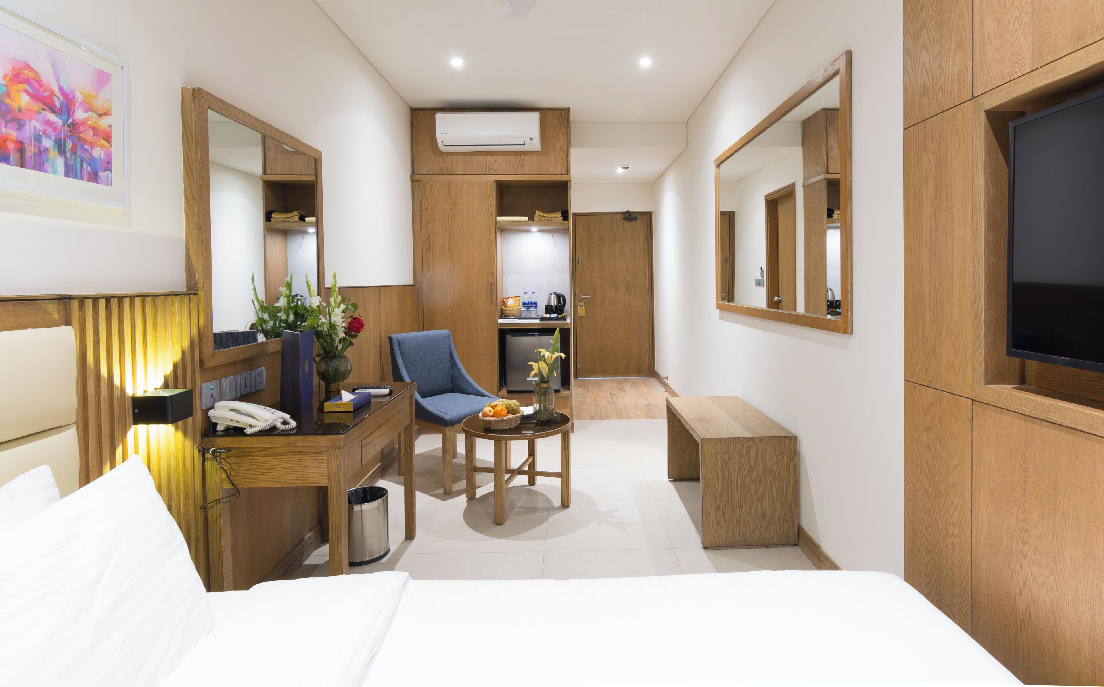A hotel room with a bed, a desk with a telephone, a chair, a small table with fruit, a trash can, a mirror, an air conditioning unit, and a television.