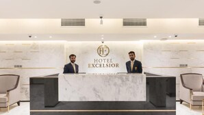Un comptoir d’accueil avec deux employés, un mur en marbre et le logo de l’hôtel EXCELSIOR.
