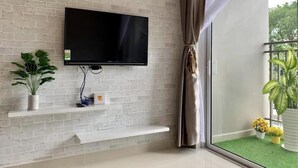 A modern living room with a flat-screen TV mounted on a white brick wall, a small shelf with decorative items, and a balcony with potted plants.