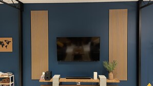 Een moderne woonkamer met een grote flatscreen-tv aan een donkerblauwe muur, omlijst door verticale houten latten. Er staat een houten bankje met een vlak oppervlak onder de tv, en een kleine plant is geplaatst aan de rechterkant.