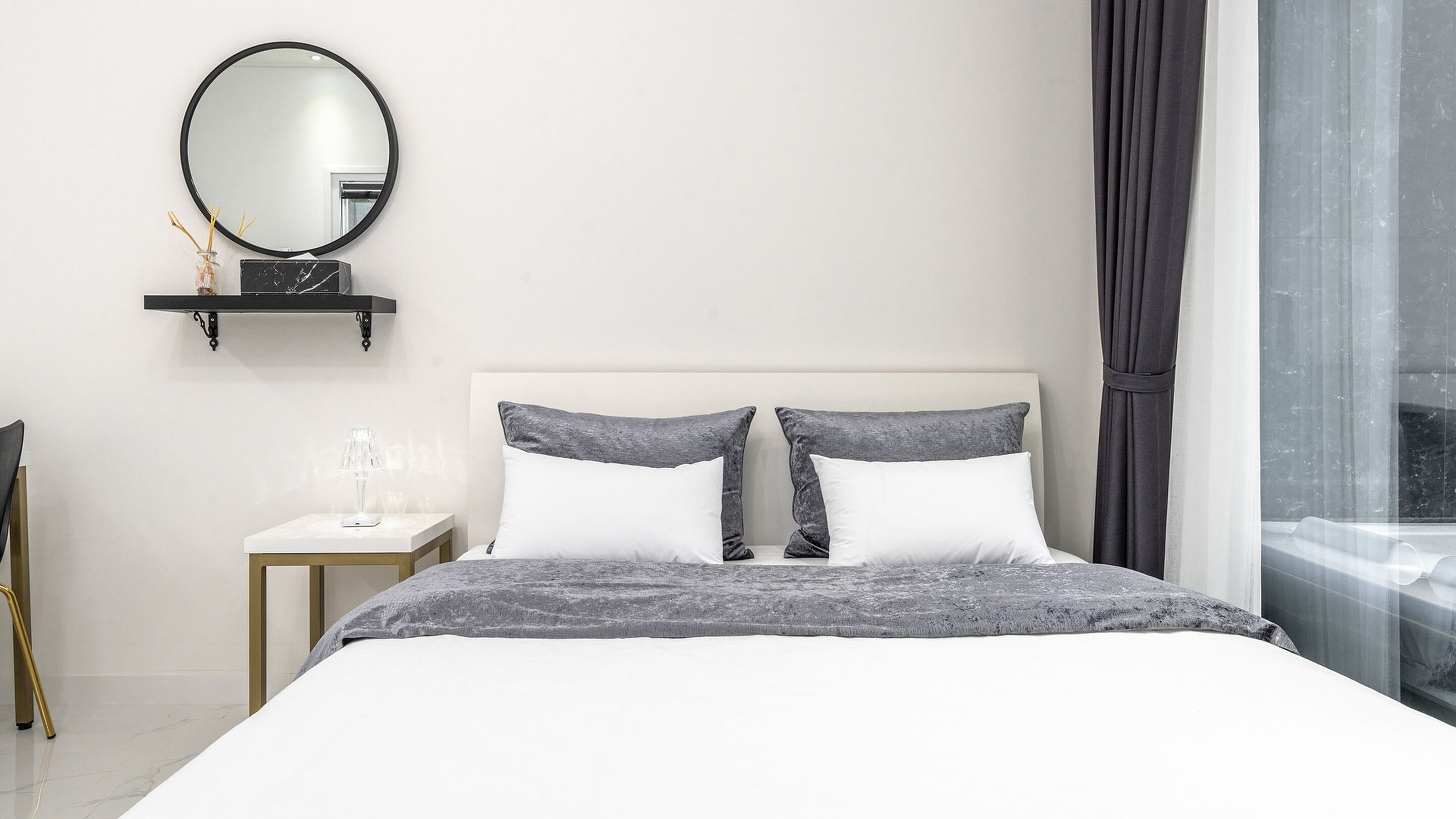 A modern bedroom with a large bed, a bedside table with a lamp, a mirror, and a window with curtains.