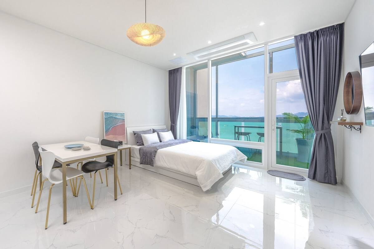 Een moderne slaapkamer met een groot bed, een eettafel en uitzicht op zee door grote ramen.