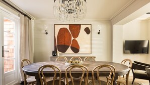A dining room with a round table, wooden chairs, a chandelier, a painting, and a TV on the wall. - Welcome to our newly renovated 4-bedroom, 2-bathroom retreat in the heart of Kensington. Warm tones, plush textures, and elegant touches create a serene space. Unique artworks and opulent accents add character throughout. Perfect getaway starts here. (London)