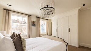 A hotel room with a large bed, a chandelier, a window with curtains, and a wardrobe. - Welcome to our newly renovated 4-bedroom, 2-bathroom retreat in the heart of Kensington. Warm tones, plush textures, and elegant touches create a serene space. Unique artworks and opulent accents add character throughout. Perfect getaway starts here. (London)