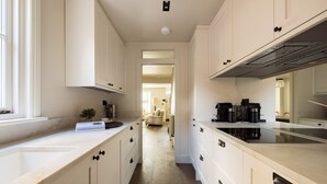 A narrow kitchen with white cabinets and a wooden floor. There is a coffee maker on the counter and a plant on the windowsill. - Welcome to our newly renovated 4-bedroom, 2-bathroom retreat in the heart of Kensington. Warm tones, plush textures, and elegant touches create a serene space. Unique artworks and opulent accents add character throughout. Perfect getaway starts here. (London)