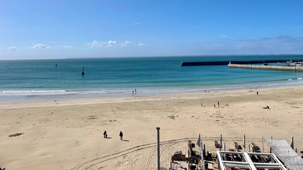 A beach with a pier extending into the water, a few people walking along the shore, and a clear sky.