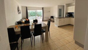 Eine moderne Küche mit weißen Schränken, eine Essecke mit Tisch und Stühlen sowie ein Gartenblick durch große Fenster.