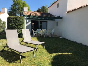 Um pátio com duas cadeiras de descanso e uma mesa, uma área externa coberta com cadeiras e um edifício branco com telhado de azulejos.