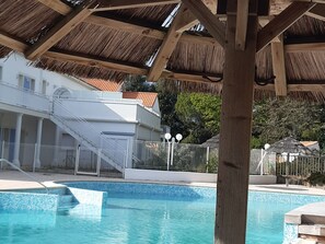 Piscina com borda azulejada, pérgola de madeira acima e um edifício branco com escadaria.